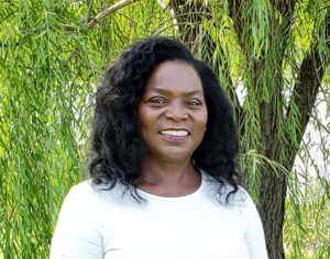 A Black woman with shoulder length black hair wearing a white long sleeve shirt smiles at camera. Green trees are in the background.