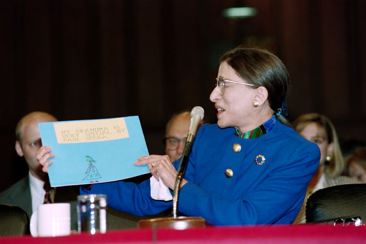 Ginsburg holds up a blue piece of paper with a child's drawing reading, 'My grandma is very special' written on it