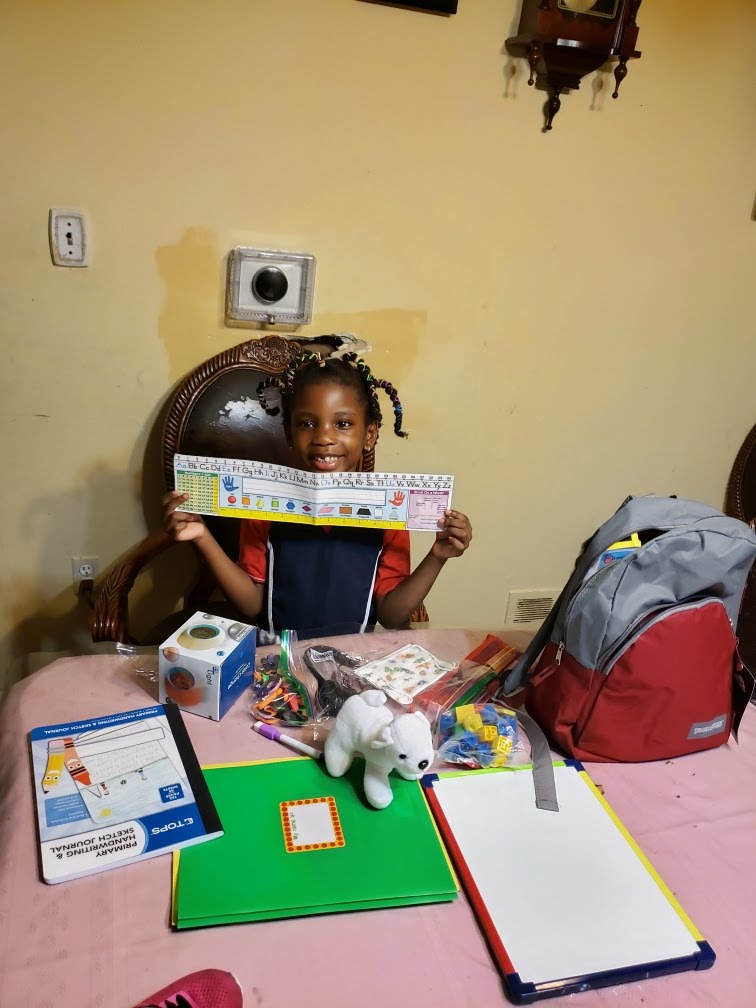 A Black child with braids smiles at the camera while holding up a paper with the alphabet. School supplies are laid out in front of her on a table with a pink table cloth.