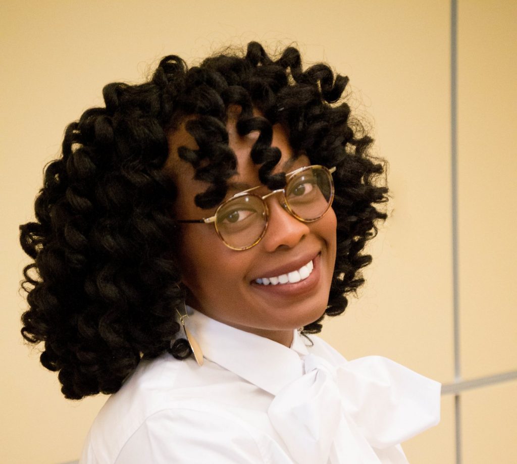 Black woman with shoulder length curly hair and glasses smiles in a portrait.