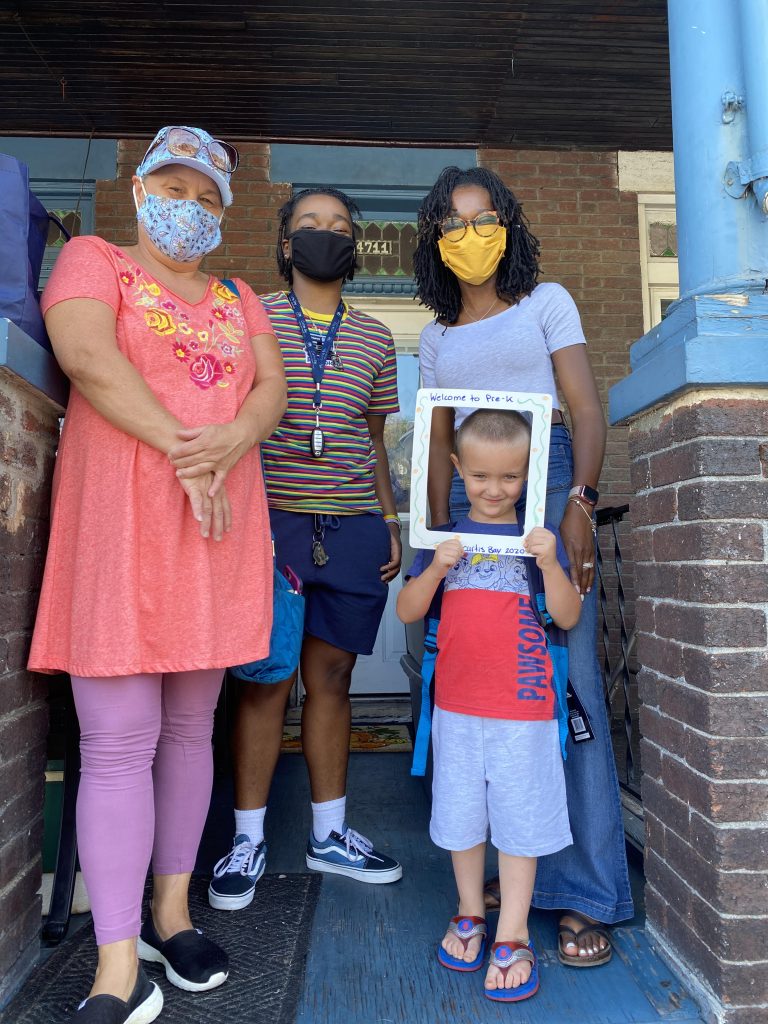 Two adults and two children stand on a porch. All wear masks.