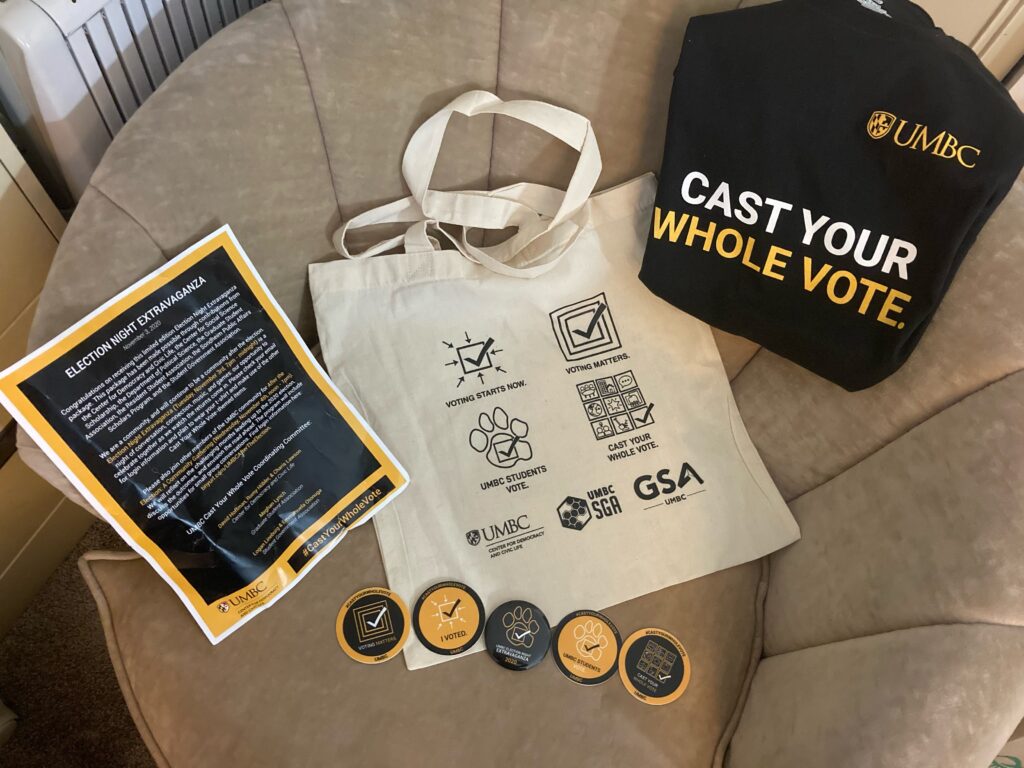A gift bag, 5 buttons, flyers, and UMBC Cast Your Whole Vote t-shirt displayed on a chair.