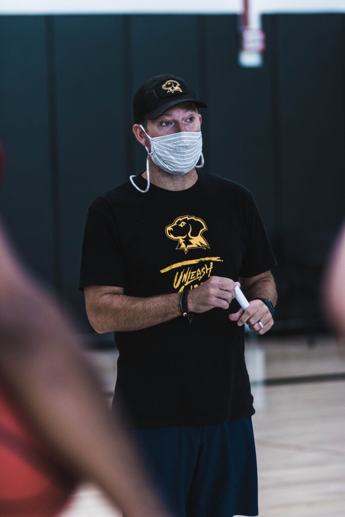 Man stands wearing black and gold Retriever hat and t-shirt, and striped cotton face mask.