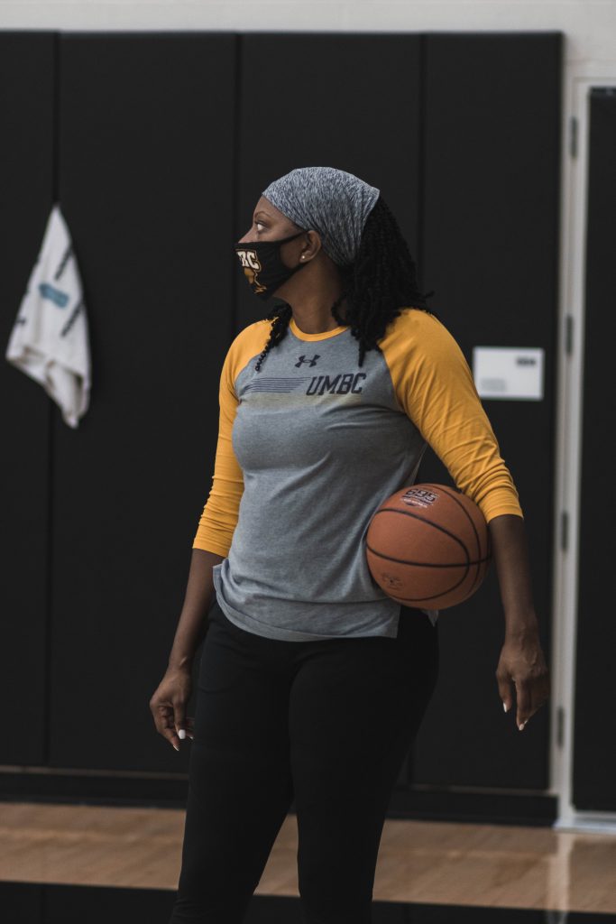 Woman wearing UMBC shirt and mask stands holding basketball
