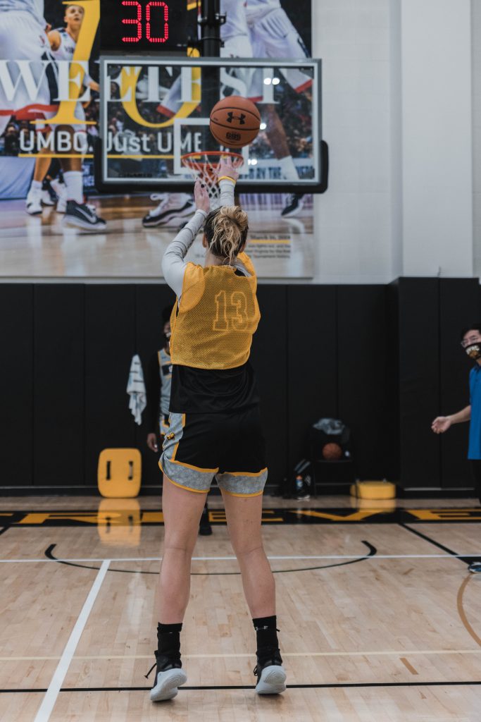 Woman seen from the back, wearing a number 13 jersey, throws a basketball into a hoop