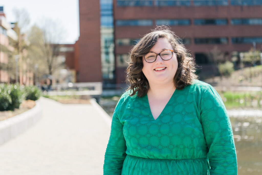 Portrait of smiling woman in long-sleeved green dress and glasses outdoors, in front of a brick building and patio.