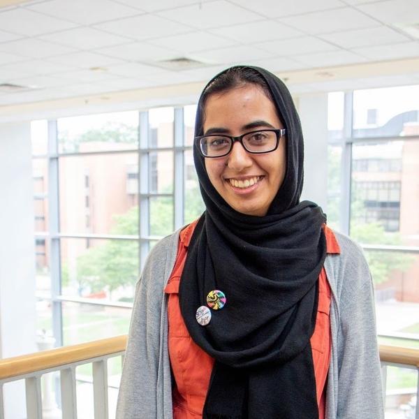 Woman in glasses, a sweater, and a shayla-style hijab with decorative buttons stands on an interior balcony in front of a window.