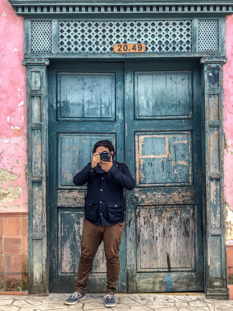 A man standing in front of a large rustic blue door, points a camera.