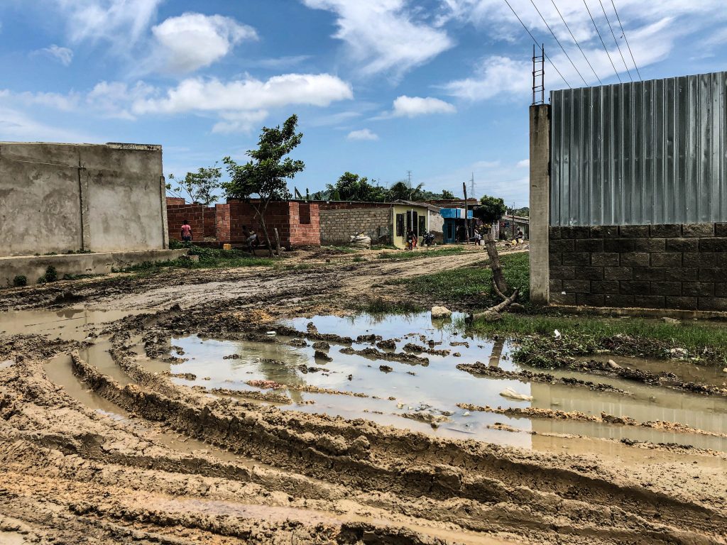 A photo of a mud filled road with tire marks left on the mud with some small brick houses in the background.