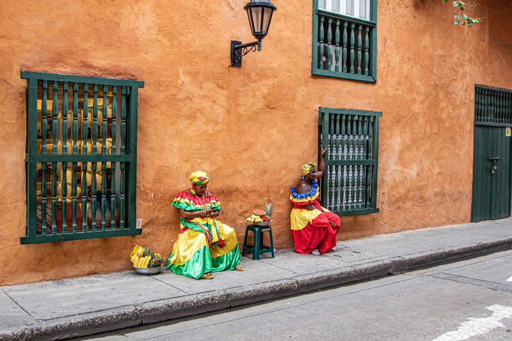Two Afro-Colombian women wearing bright long colorful skirts, shirts, and head coverings sit with two bowls of fruit next to them as they lean on the orange wall of a building.