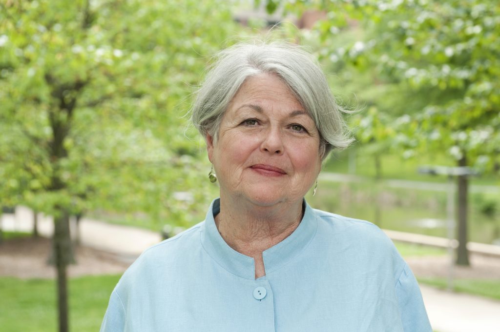 A woman with short grey hair wearing a light blue blouse and earrings stands in front of a group of green trees and smiles at the camera.