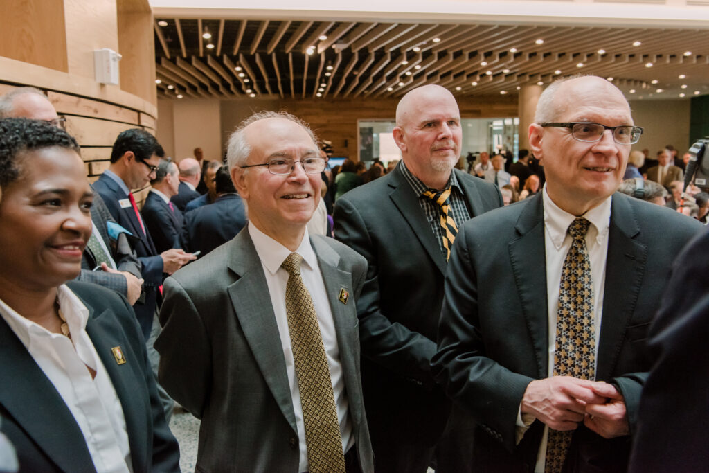 Four people in business attire conversing in a large open atrium, many more people behind them