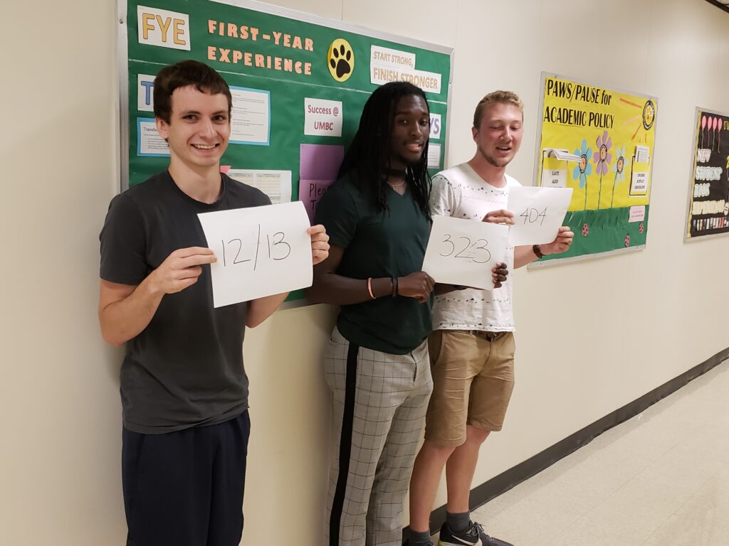 Three young men stand in a hallway holding up white pieces of paper with numbers written in black. There are colorful bulletin boards behind them.