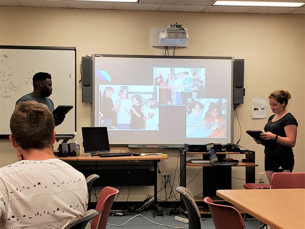 A young man and a young woman holding black tablets stand in front of a room and flank a large screen displaying three photos of groups of students.