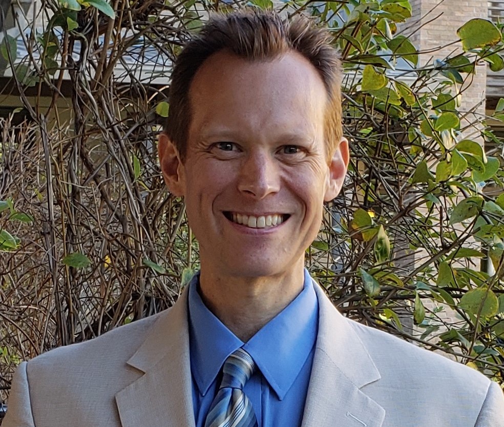 A man with short brown hair and wearing a light grey dress jacket, blue dress shirt, and gold and blue striped tie smiles at the camera.