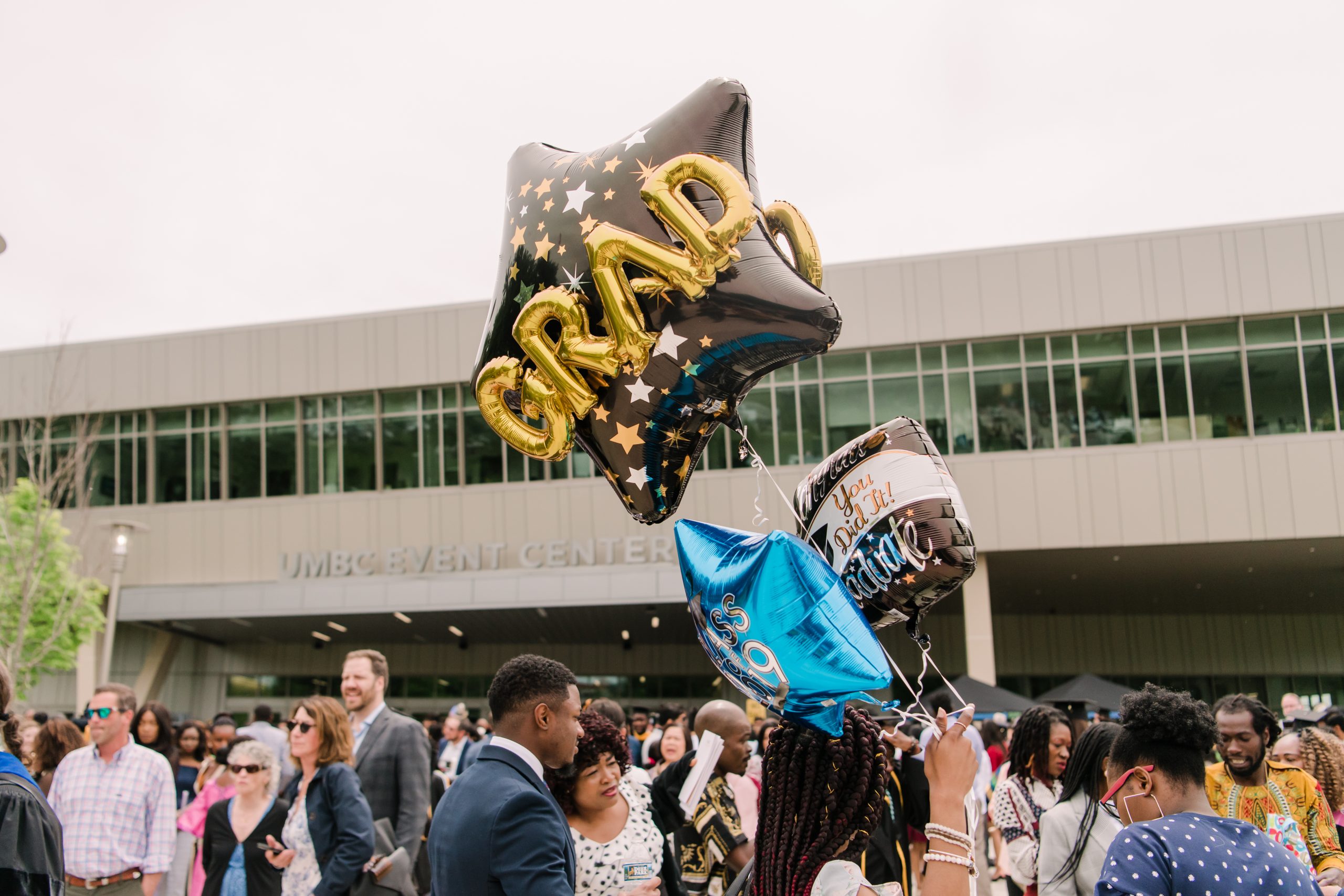 Star-shaped graduation balloons float in front of the UMBC event center. A crowd of people are outside waiting for graduation commencement to begin.