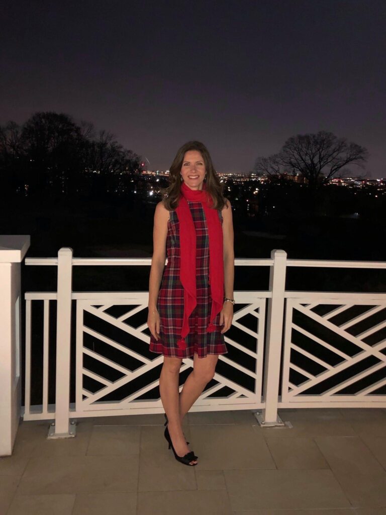 White woman in dress and scarf stands outdoors in the evening, next to a white fence.