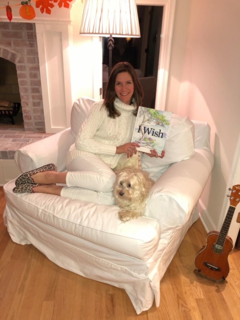 White woman with long brown hair sits on a white couch, next to a small dog, holding a book titled 