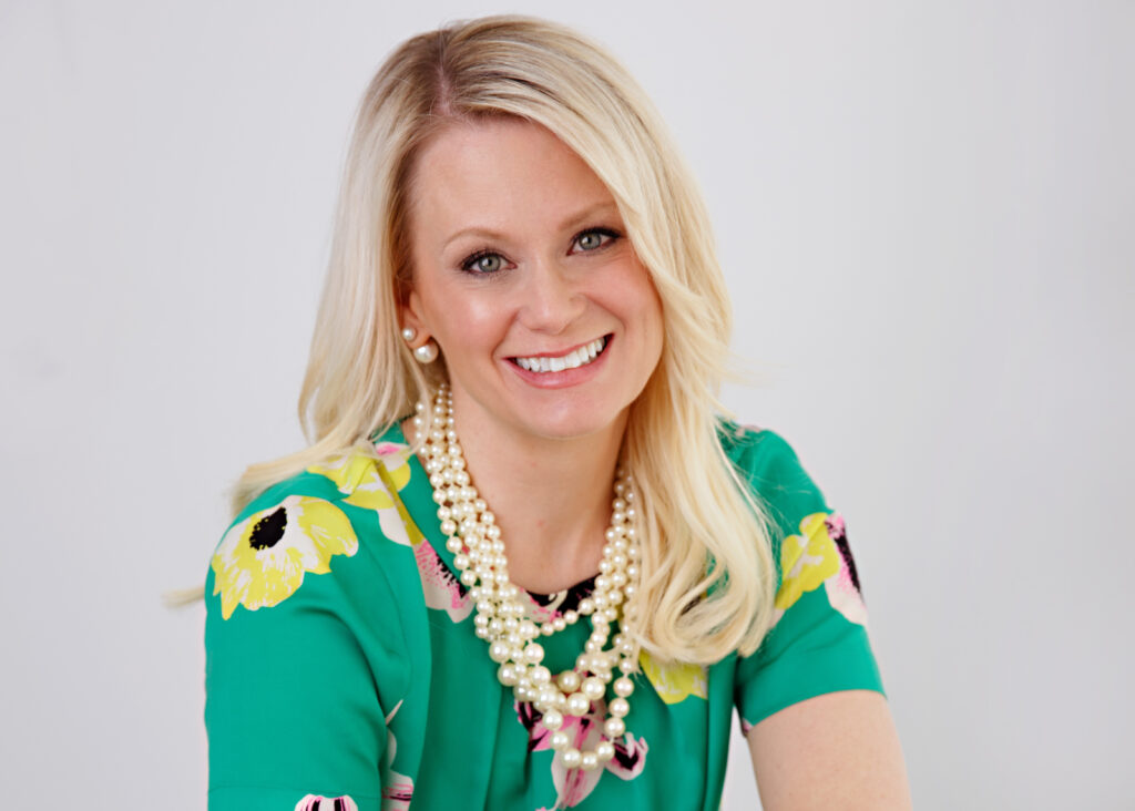 A blond woman wearing strings of pearls and a green blouse with yellow flowers smiles at the camera.