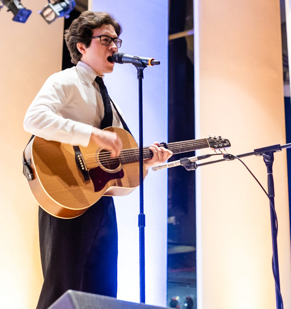 Young man in dress shirt and tie sings into a mic while playing guitar.
