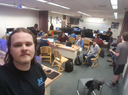 Young white man in black t-shirt looks at the camera, while several other students are seated and standing in the background, around tables and laptops.