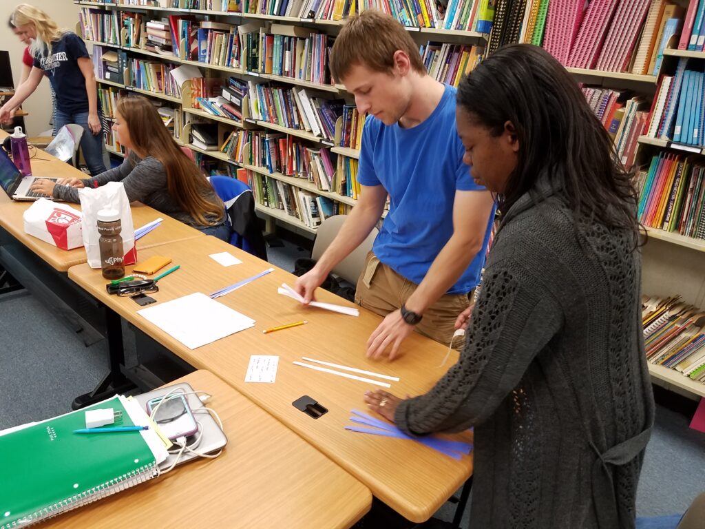 Three young women and one young man work around two wodden rectangular tables with white and blue strips of paper. There is a row of shelves behind them. 