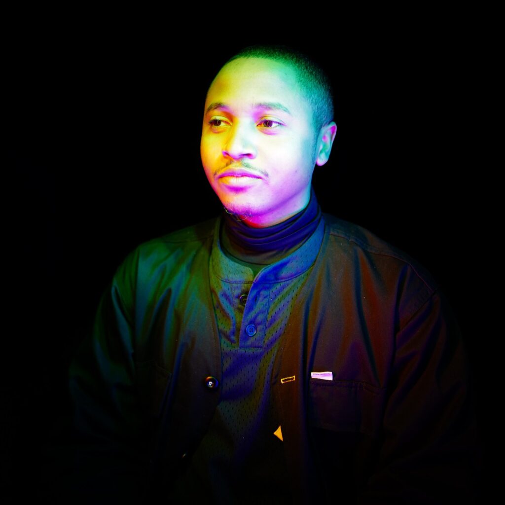 Portrait of a young black man lit by multi-colored neon lights. He wears a buttoned shirt and jacket.