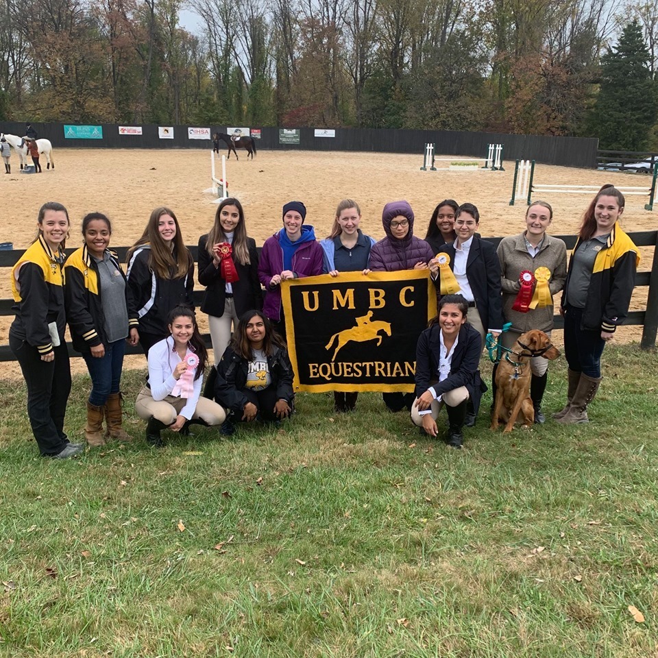 A group of fourteen young women stand together holding a gold and black banner that says UMBC Equestrian. Behind them is a fenced-in dirt area with horses.