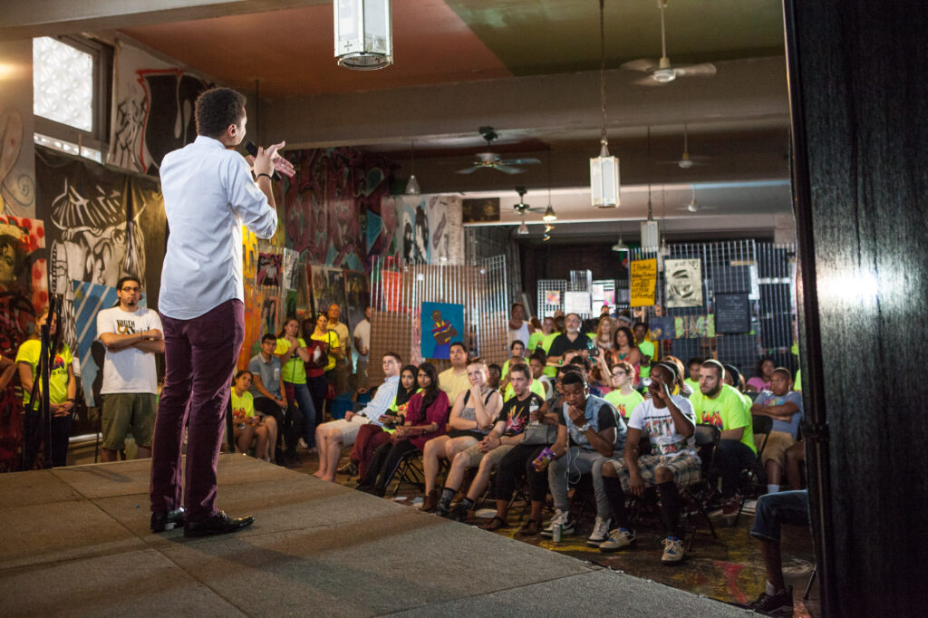 Young man stands on a stage speaking to a crowd of seated young people in a small rooms. Paintings and posters hang on the walls.