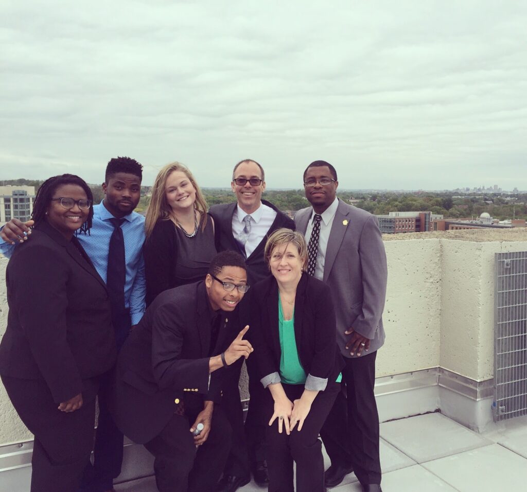 Seven people stand on a rooftop, smiling for a portrait. They wear professional clothing, including blazers.
