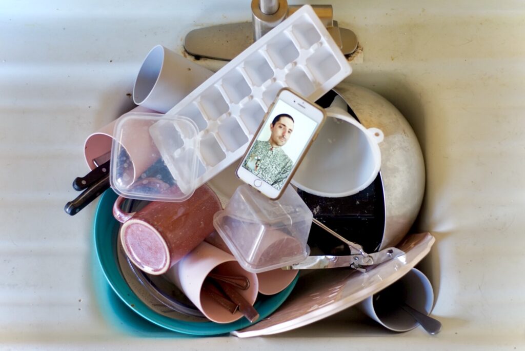 iPhone displaying a Facetime video sits in a sink with a pile of dirty dishes.
