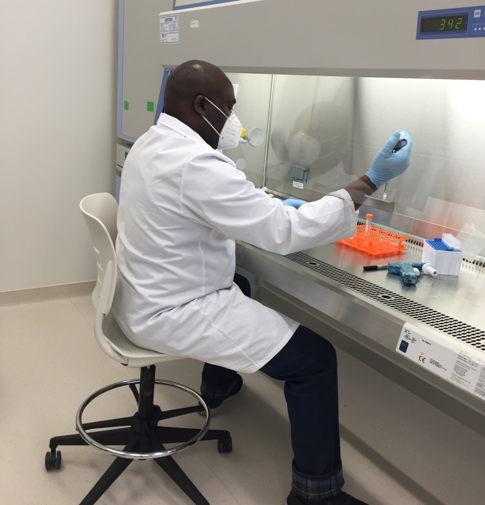 Man sits on a stool at a fume hood, wearing a lab coat and pipetting