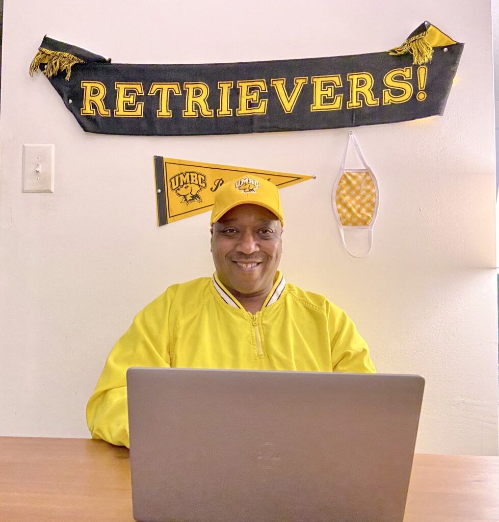 A man sits behind a laptop with a gold shirt on and a banner that says Retrievers! behind him