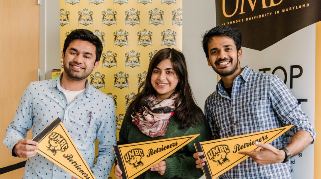 Three young adults smile at camera, holding pennants that read UMBC Retrievers