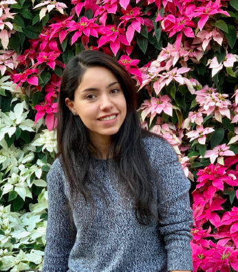 A young woman with long black hair wears a grey sweater smiles at the camera with a wall of pink and white flowers.