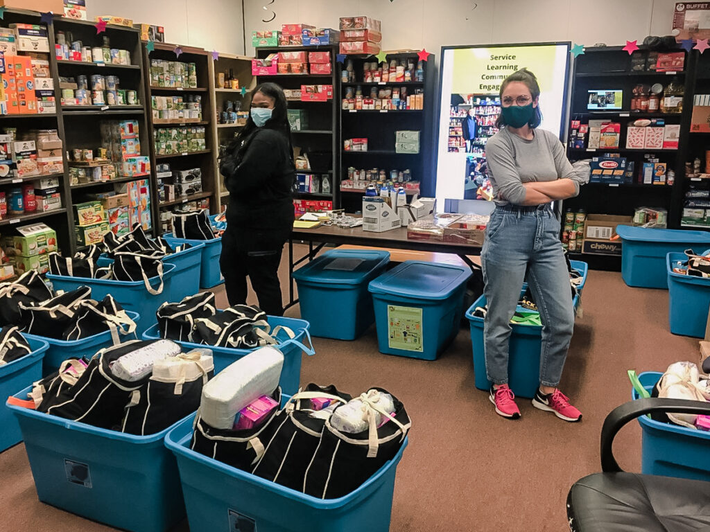 Two young women wearing blue face masks stand facing away from each other while smiling at the camera in a room lined with shelves filled with cans of food. On the floor around them are large blue tubs with black and white bags filled with a variety of objects.