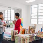 One woman wearing a bright red dress is speaking with another woman holding a baby. They are standing in front of a table stacked with children's story book in white room with windows lining the wall behind them.