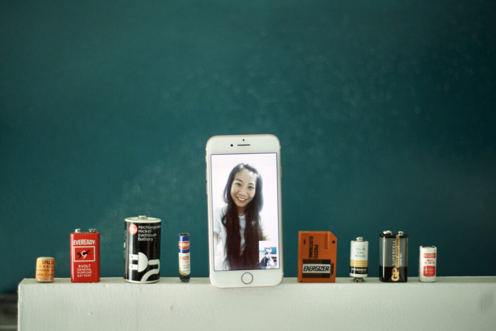 iPhone displaying a Facetime video of an Asian woman stands in the middle of different types of batteries