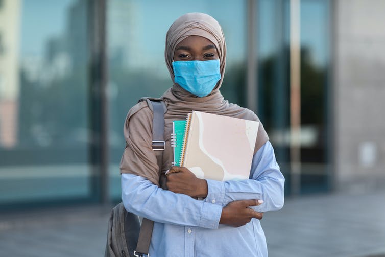 Black muslim female student in mask with workbooks outdoors