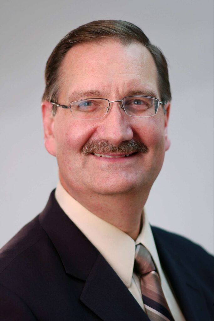 Portrait of middle aged white man with mustache and wire-framed glasses. He wears a dark suit with striped tie.