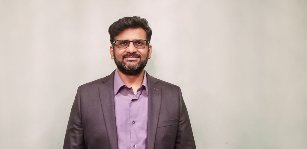 Portrait of a middle-aged South Asian man. He has a beard and wavy hair, and wears wire-framed glasses and a suit.