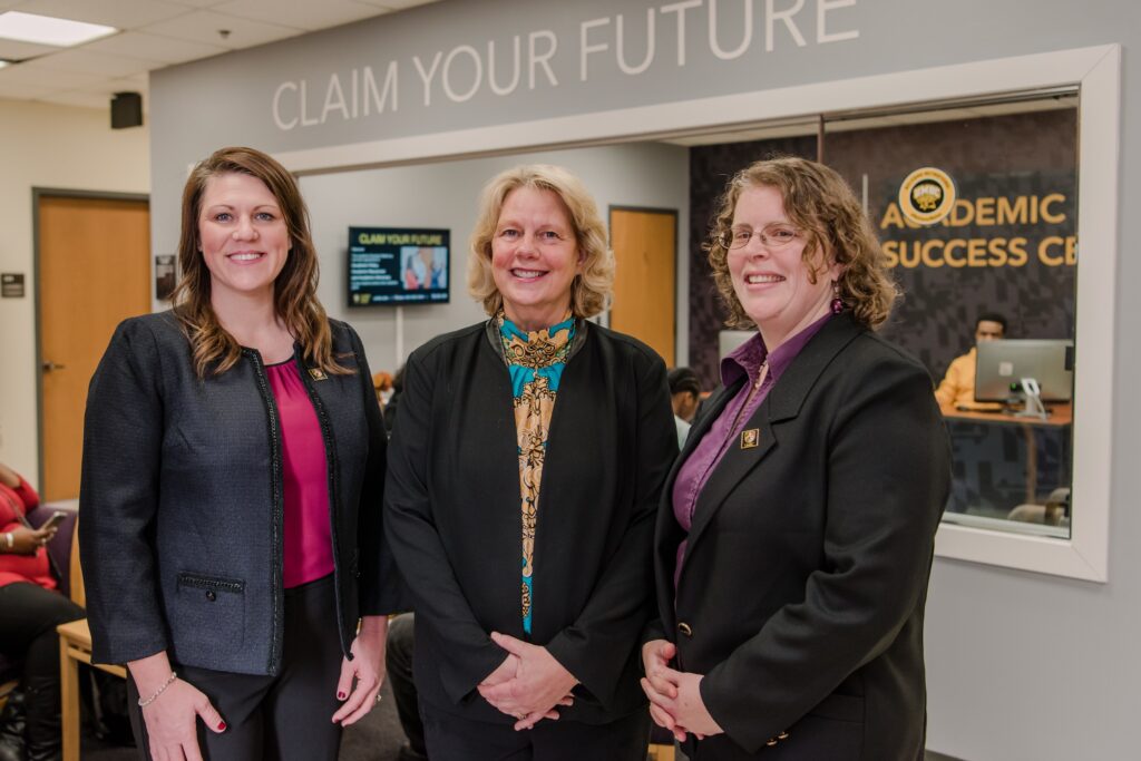 Three white woman in black blazers and dress shirts stand in an office lobby. Behind them a sign reads 