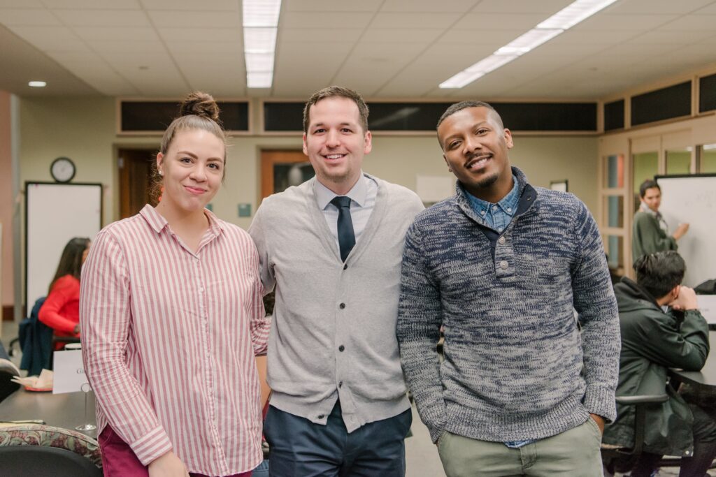 Three young professionals stand together, wearing collared shirts and sweaters.