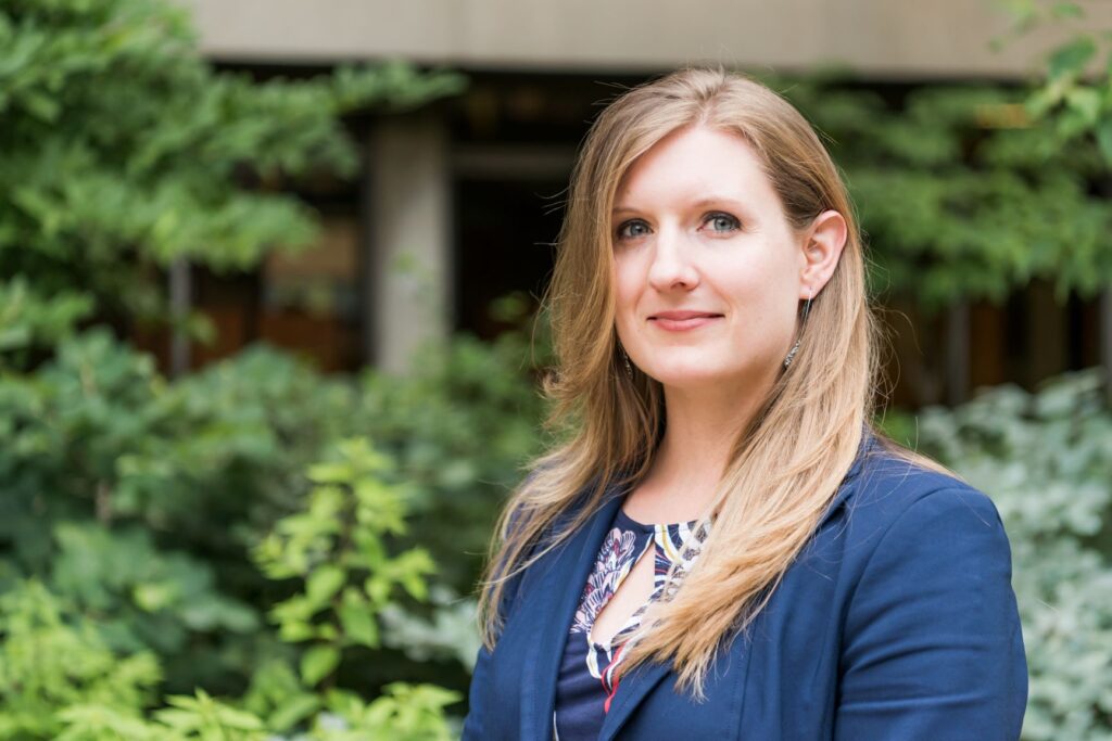 Outdoor portrait of a middle-aged white woman with long blonde hair. She wears a navy suit jacket and looks at the camera with a positive but serious expression. Plants are in the background.