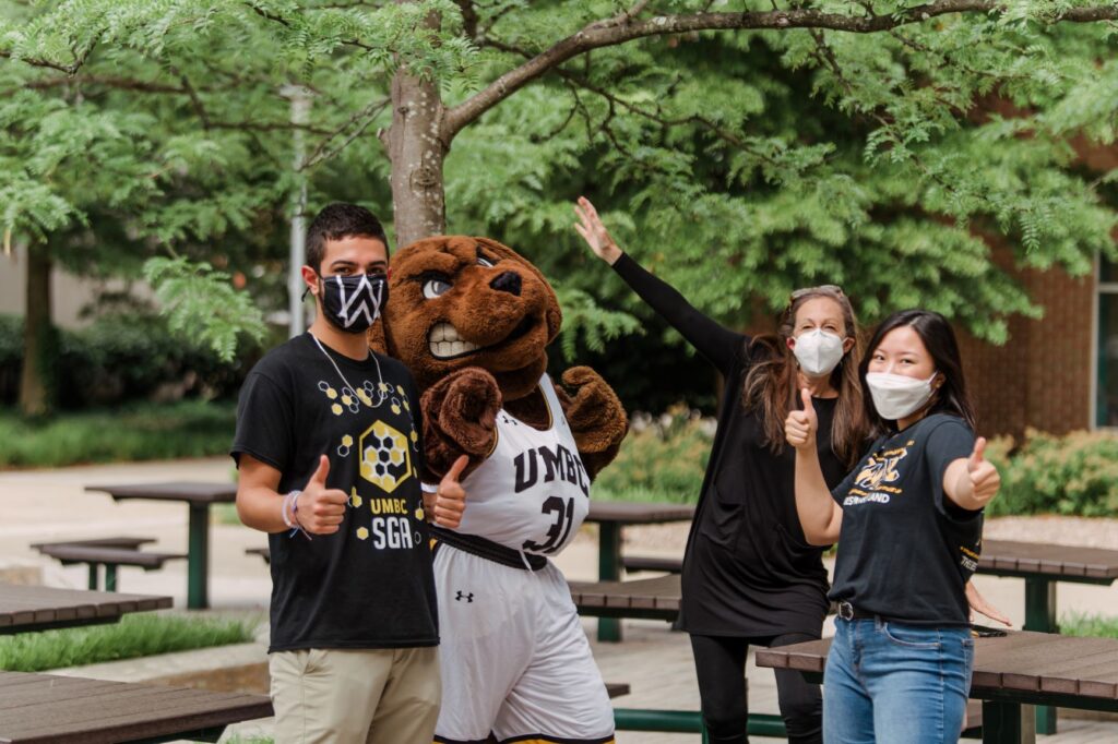 Two young adults and one middle-aged adults stand outside, giving thumbs up signs. A person wearing a Retriever mascot in UMBC jersey joins them.