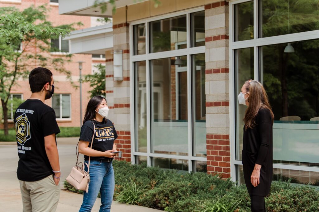 Three people stand outside a building, all wearing masks. Two wear black and gold t-shirts, with one reading 