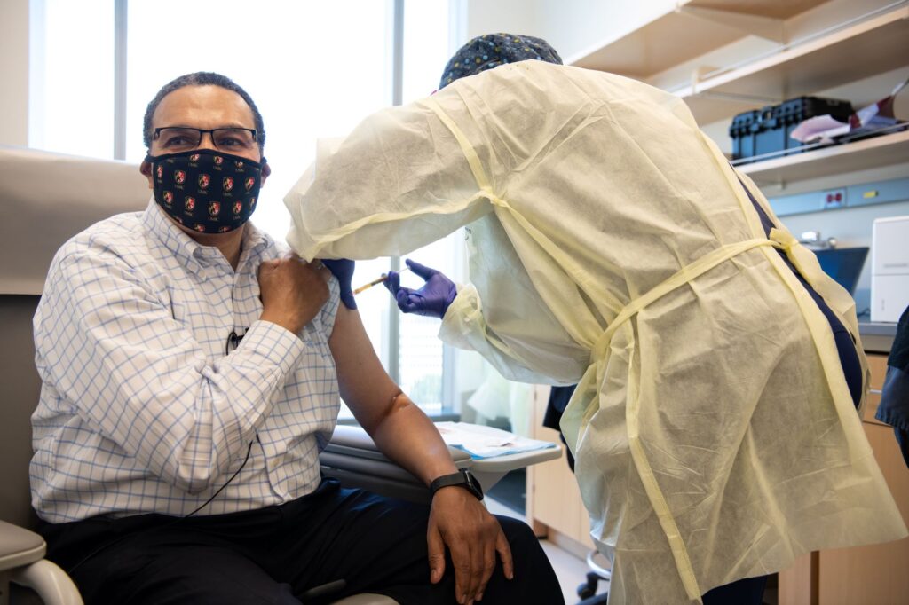 Middle-aged black man with glasses and a face mask receives a shot from a medical professional wearing protective gear. He wears a blue and white dress shirt and dark slacks.