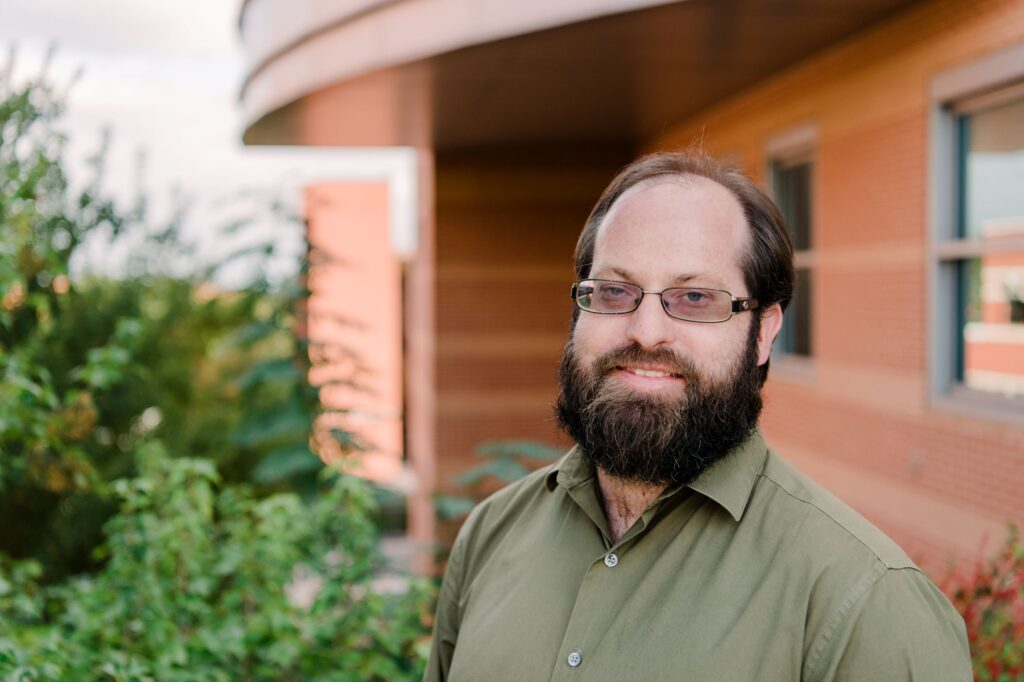Portrait of a middle-aged white man with full beard. He wears a green dress shirt and wire-framed glasses. He stands in front of a brick building and plants.