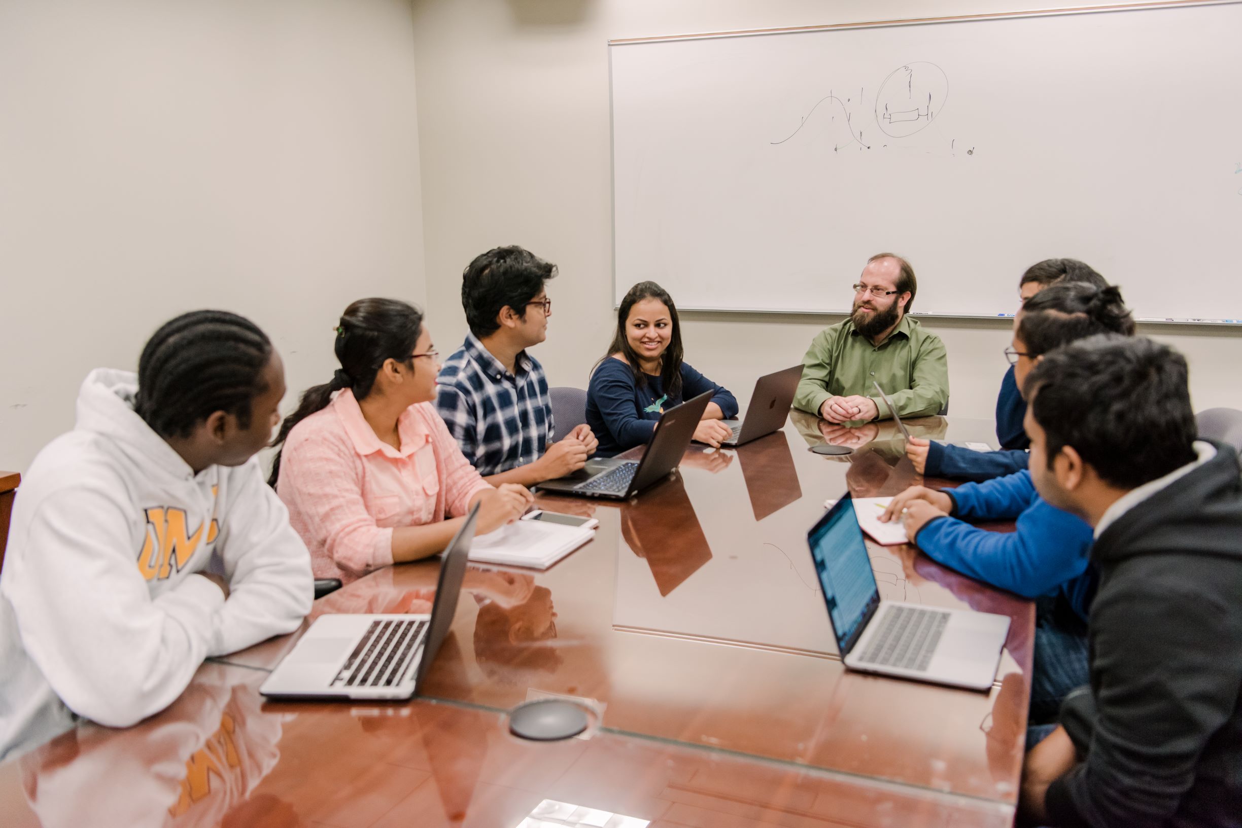 Seven students and a professor sit around a conference table. Five students have open laptops and two are taking notes on paper.