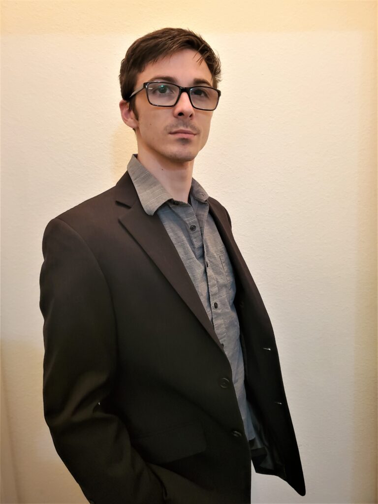 A young man with short brown hair wearing dark rimmed glasses and a brown dress shirt and jacket stands in front of a beige wall and looks into the distance.
